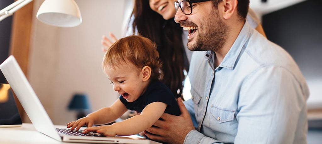 A smiling man holds a happy toddler in a dark blue shirt as the child presses keys on a laptop, while a joyful woman looks on in the background.