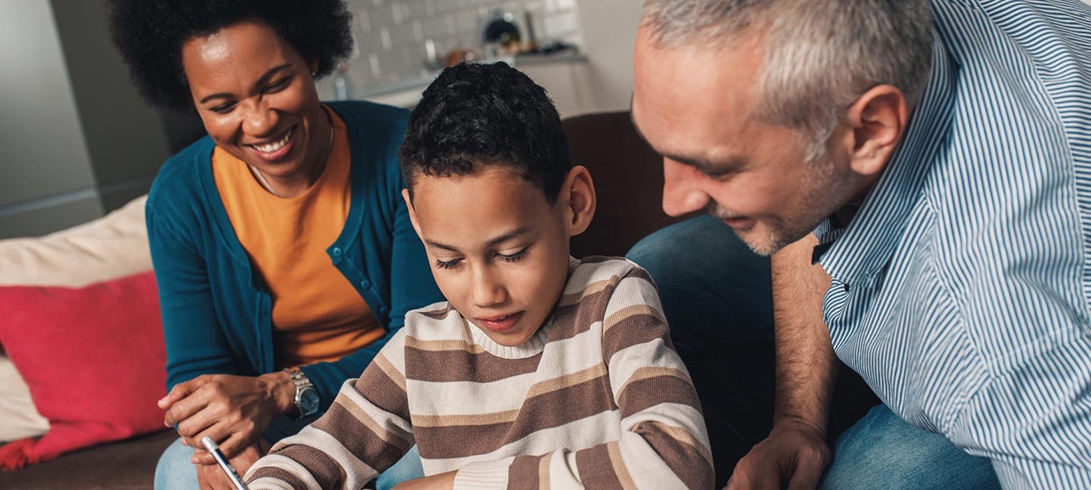 A mother, father, and son of mixed ethnicity are sitting close together, all facing the boy's sketchpad. The boy is drawing with a pencil. The parents are watching the boy with a kind smile. The family is sitting indoors on a brown sofa.