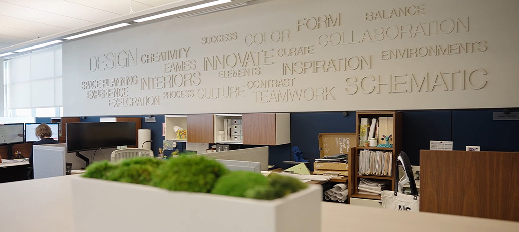 Modern open-concept office workspace with cubicles, shelving, and a planter in the foreground.