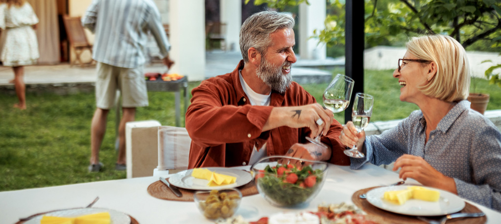 Two people sit at a dining table, raising glasses of white wine during a relaxed meal.