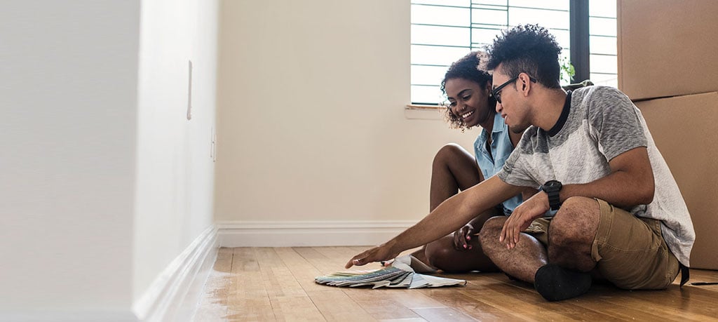 A young couple sits on the floor of their new home, contemplating paint swatches. The room is a jumble of moving boxes and a lone backpack. The woman grins as the man gestures at the color options. A window peeks out in the background.
