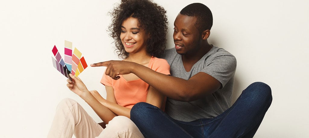 A smiling African American couple sitting on the floor in front of a white wall, reviewing paint color swatches. The man is pointing to a color on the swatches.