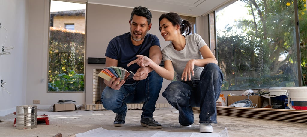 Happy couple crouching on an unfinished floor in their home, examining a fan of colorful paint swatches together during a renovation project, with large windows and construction materials visible in the background.