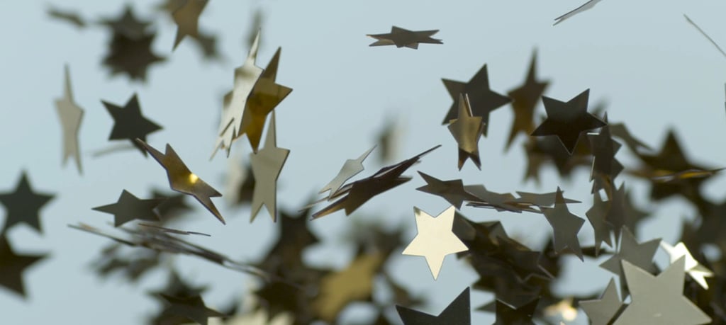 Metallic gold and silver star confetti pieces suspended in mid-air against a soft blue-gray background, creating a celebratory effect