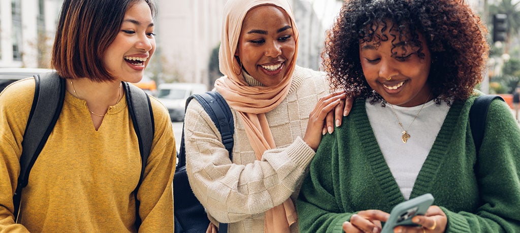 Three people walk side by side on a city street, wearing casual clothing and backpacks, with one person holding a phone.