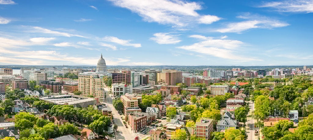 A wide, high-angle shot captures the cityscape of Madison, Wisconsin, under a bright, partly cloudy sky. The Wisconsin State Capitol building, topped with a distinctive dome, stands prominently amidst a mix of modern high-rise buildings and traditional low-rise structures. Lush green trees fill the spaces between buildings, creating a vibrant, green urban landscape. The perspective gives a comprehensive view of the city's layout and architectural diversity, emphasizing its blend of natural beauty and urban development.
