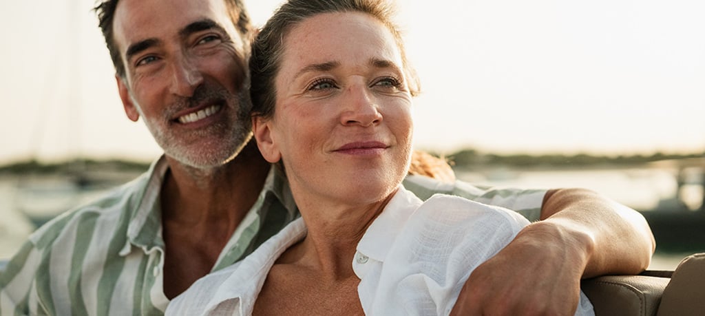 Two people sit close together on a boat, with one person’s arm resting along the back of the seat as water and shoreline appear in the background.
