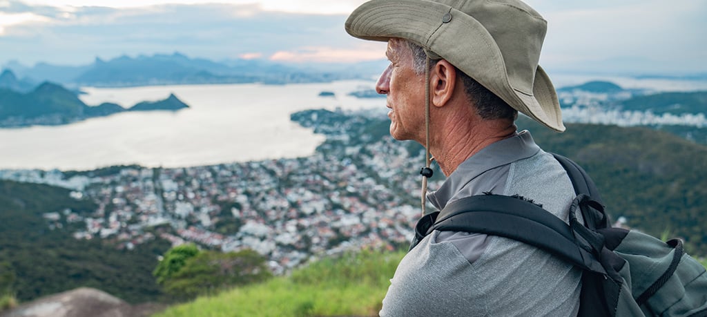 A hiker with a backpack stands on a grassy overlook, taking in a sweeping view of a coastal town and water below.
