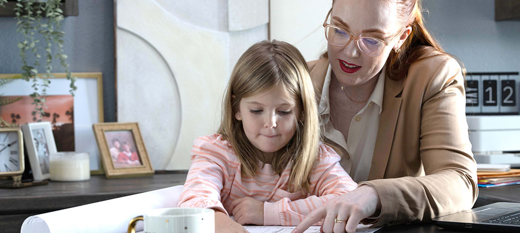 An adult in a tan blazer and glasses points to a document on a desk, while a young child in a striped shirt looks intently at it. They are in what appears to be a home office setting, with framed photos and a plant in the background.