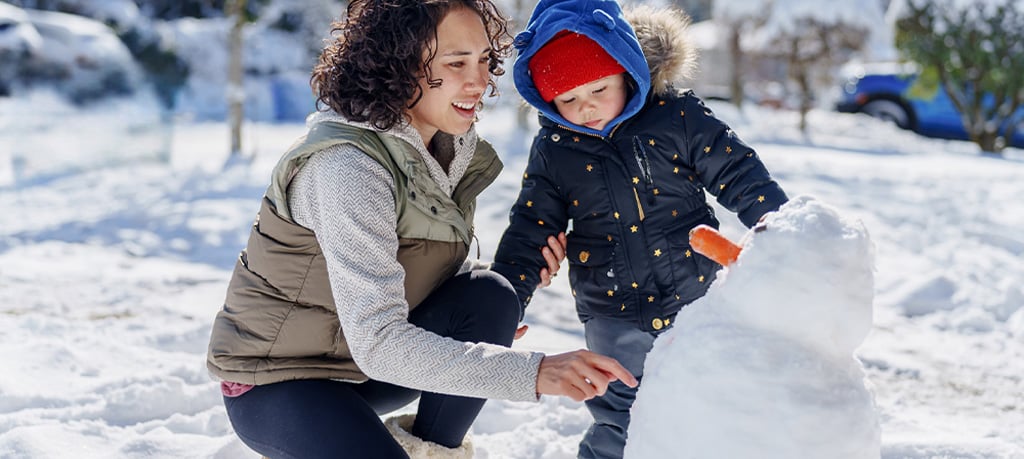 A woman and young child building a snowman together in a snowy outdoor setting. The child wears a navy winter coat with star patterns and orange mittens, placing their hand on the snowman while the woman crouches beside them, both smiling.
