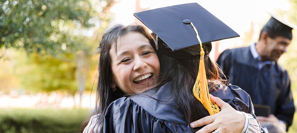 A beaming woman hugs a graduate in a cap and gown in this eye-level shot. The woman, with dark hair and a bright smile, is the primary focus, her arms wrapped around the graduate. The graduate wears a traditional black mortarboard cap with a gold tassel. In the soft-focus background, another person in graduation attire is visible, holding a phone and smiling. The outdoor setting has trees and a building, bathed in soft sunlight.