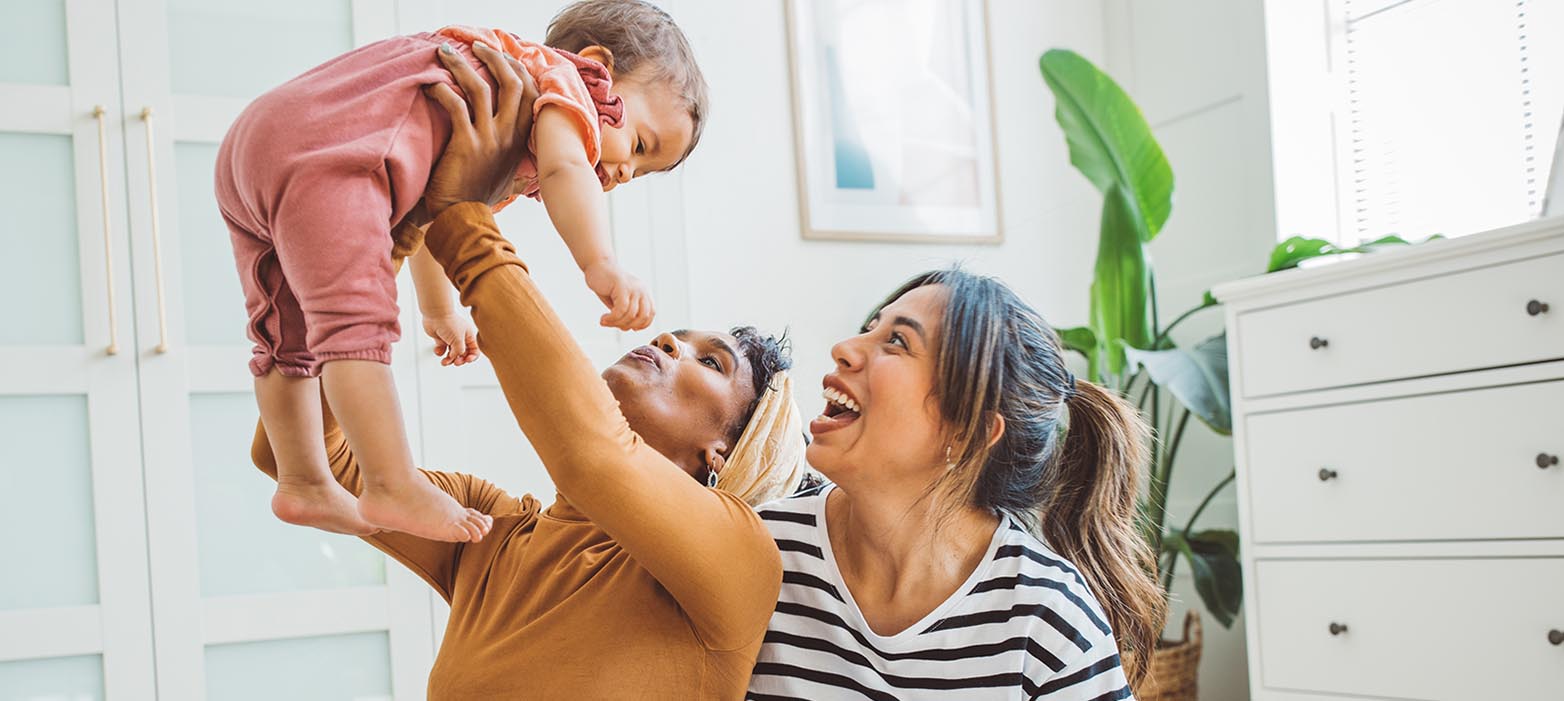 A woman holds a baby above her head while another woman sits next to her, looking up at the baby and smiling. The woman holding the baby wears a brown long-sleeved shirt and blue jeans and has her arms extended straight up to hold the baby. The baby wears a pink onesie and is looking down at the woman holding her. The woman next to them wears a black-and-white striped shirt and blue jeans and has her mouth open in a smile. They are all sitting on a colorful rug in a room with white walls and a white dresser.