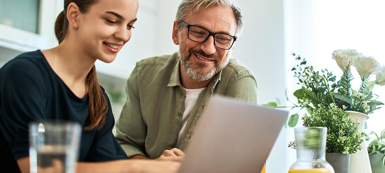 A smiling young woman and an older man, possibly her father, are sitting at a table looking at a laptop screen. The woman is typing on the laptop, and the man is watching the screen with a smile. There are drinks and plants on the table.