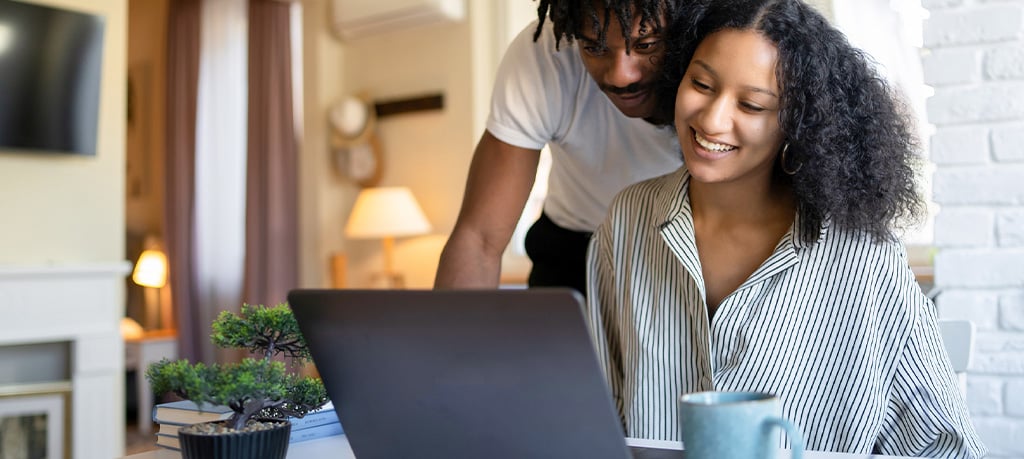 Two people smiling while working together on a laptop at home, with a small bonsai tree and coffee mug on the table, in a warm and inviting living room setting