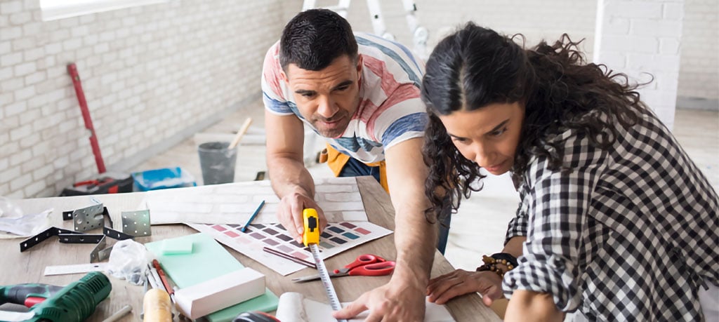 A man and woman work together on a home renovation, carefully measuring wallpaper and comparing color swatches. Tools and building materials are visible on the table around them.