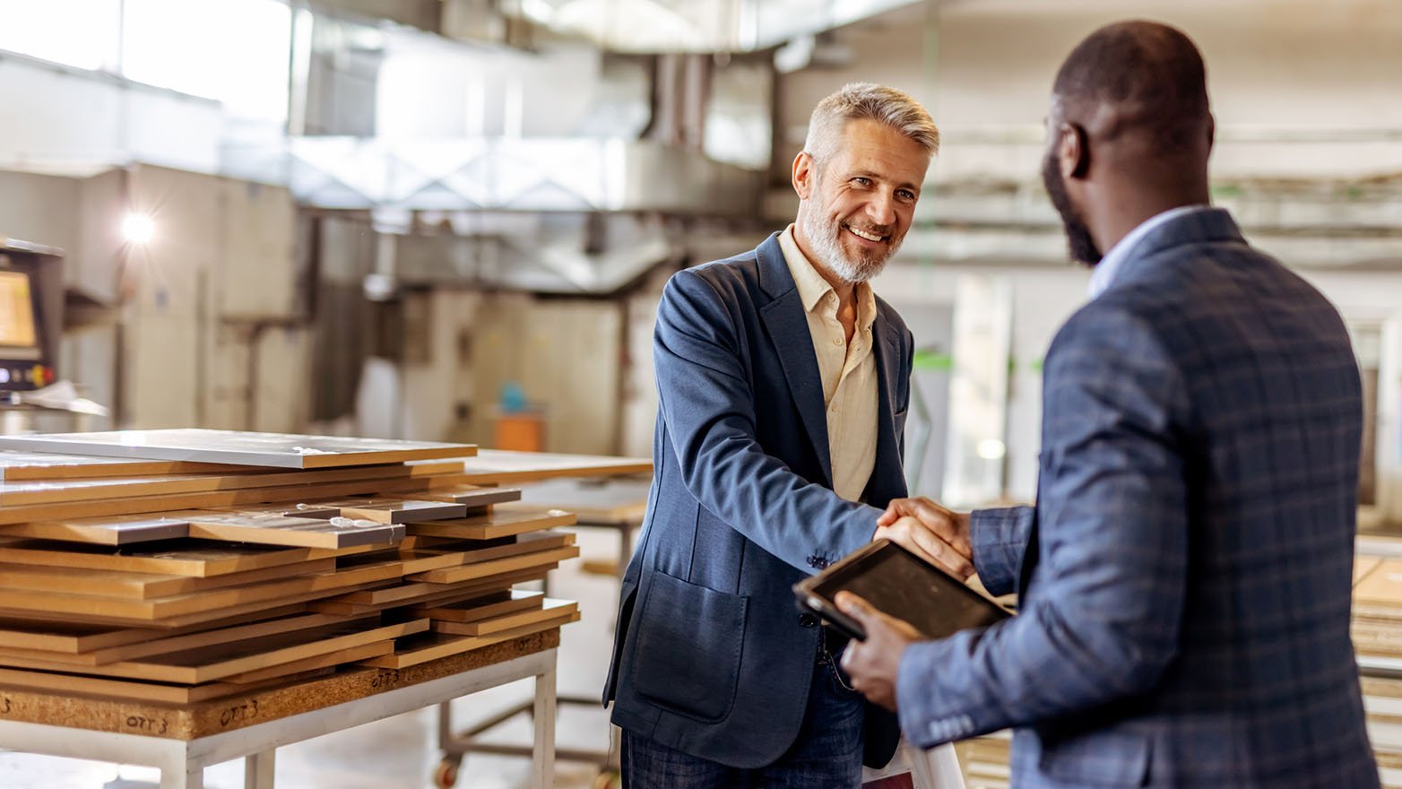 Two men, one in a blue blazer and the other in a plaid jacket, shake hands and smile inside a factory.