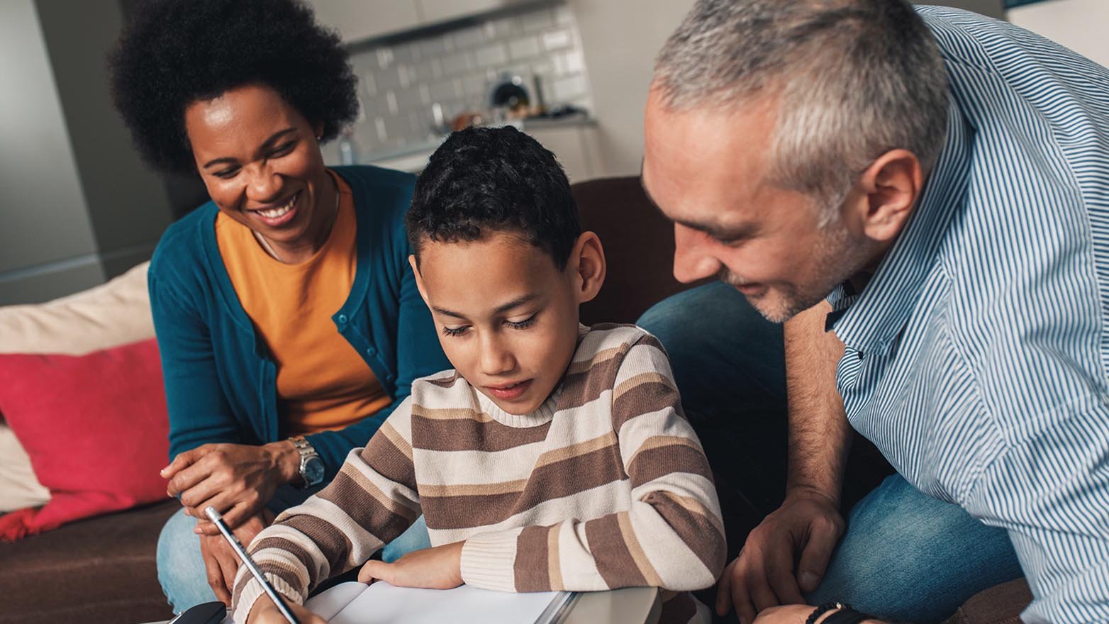 A mother, father, and son of mixed ethnicity are sitting close together, all facing the boy's sketchpad. The boy is drawing with a pencil. The parents are watching the boy with a kind smile. The family is sitting indoors on a brown sofa.