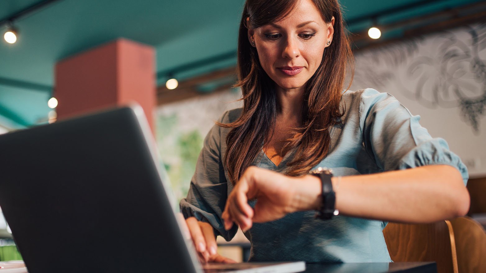 A person seated at a café table works on an open laptop while glancing at a wristwatch, with warm indoor lighting, a teal ceiling, wall art, and seating visible in the background.