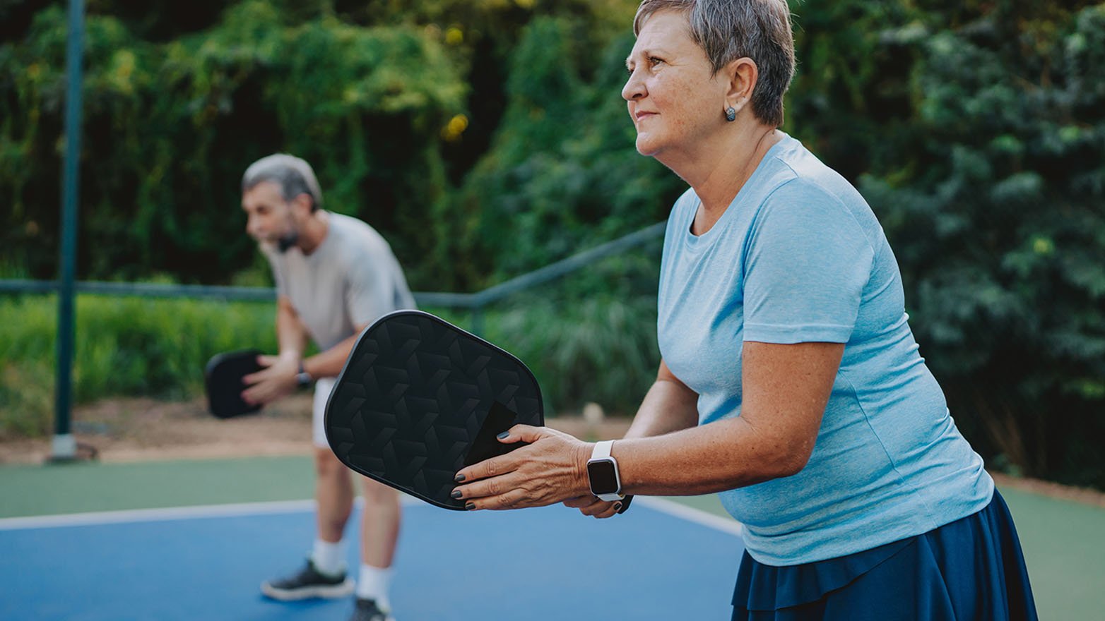 Two people play pickleball on an outdoor court, with one person holding a paddle and preparing to return the ball.