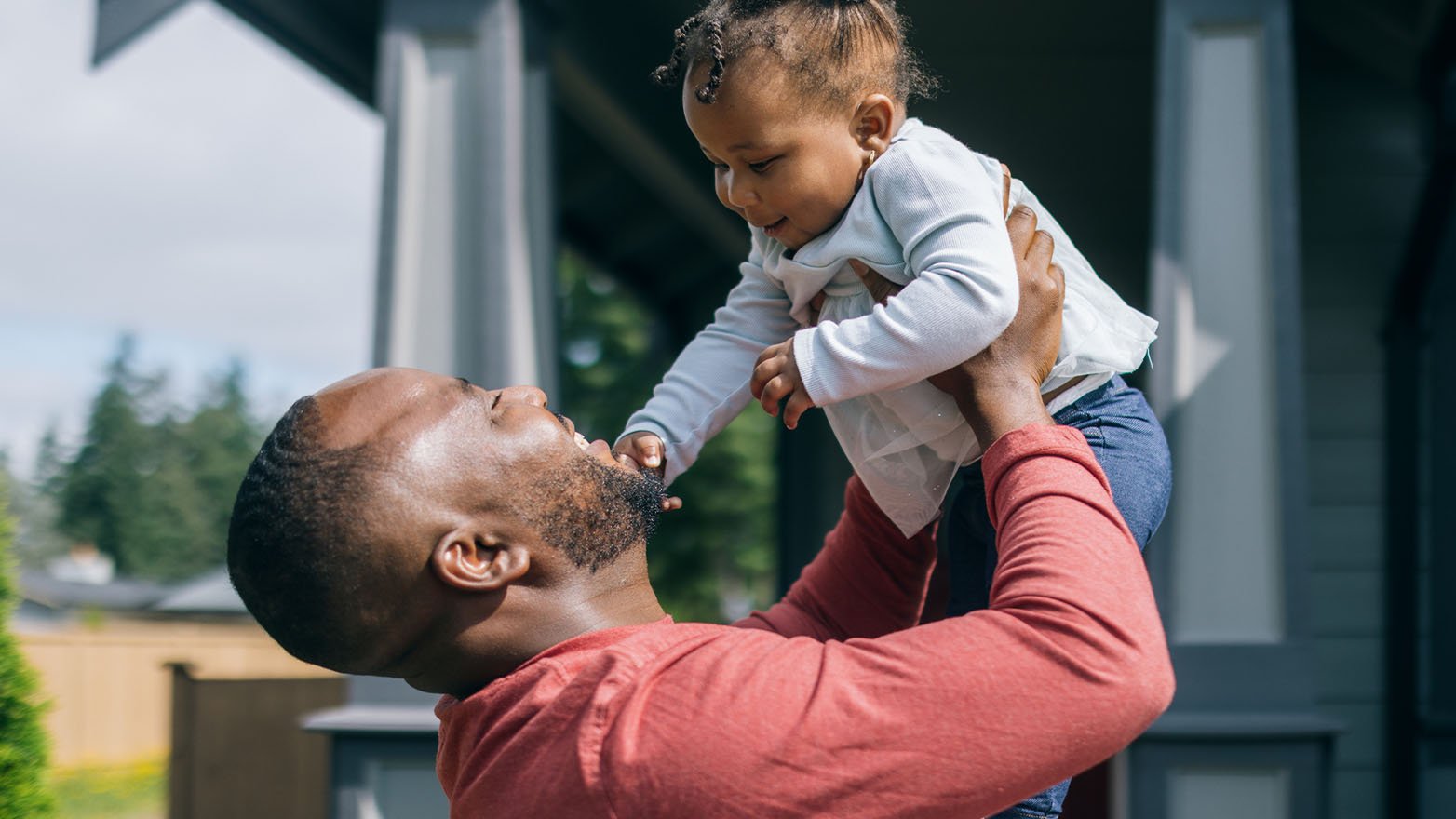 Joyful moment between father and child as dad lifts his smiling baby high overhead in front of their house. The father wears a red long-sleeve shirt and looks up at the baby with delight. The baby, dressed in a light blue outfit with hair styled in small buns, smiles down at their father against a backdrop of the home's dark exterior.