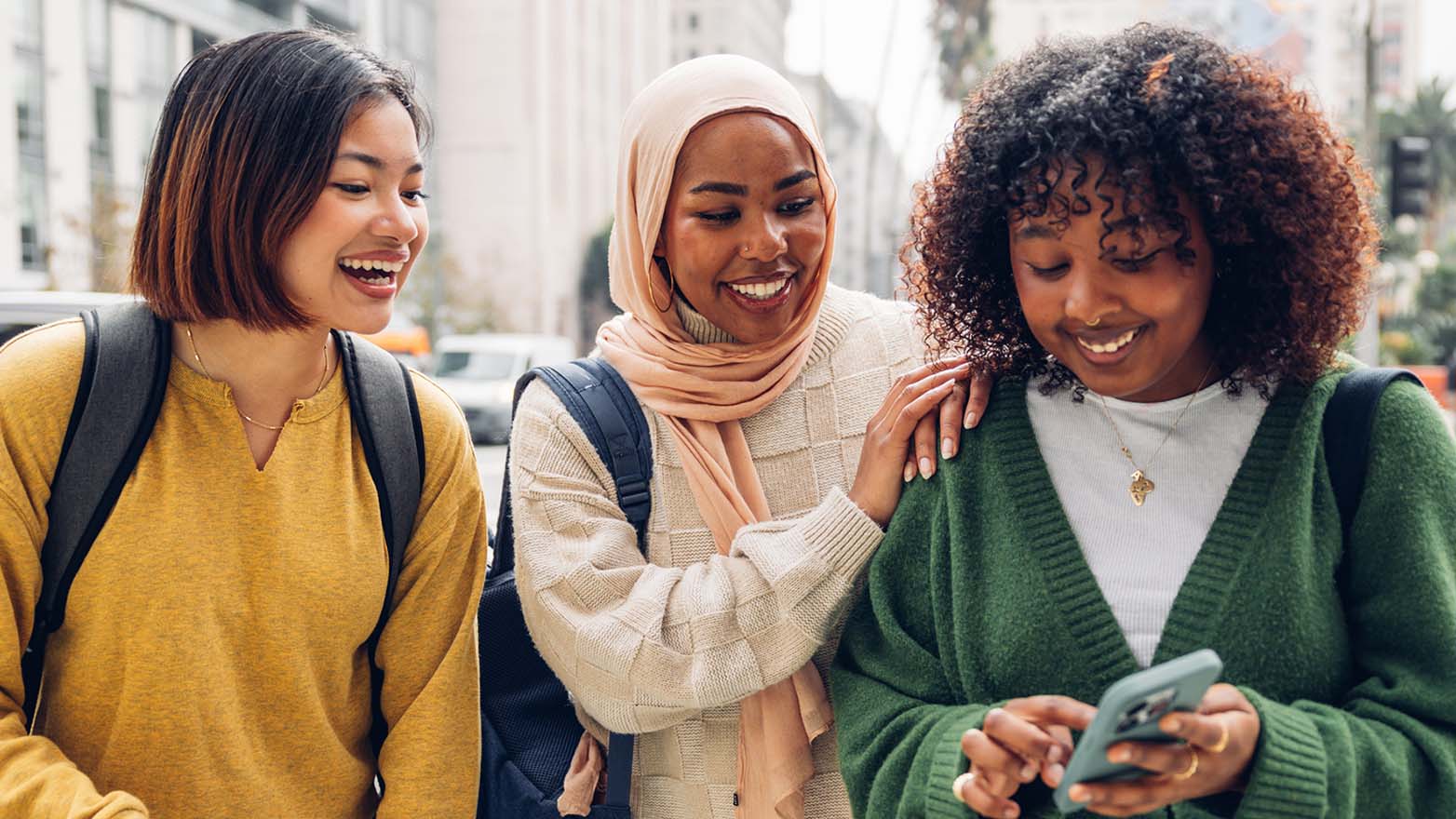 Three people walk side by side on a city street, wearing casual clothing and backpacks, with one person holding a phone.