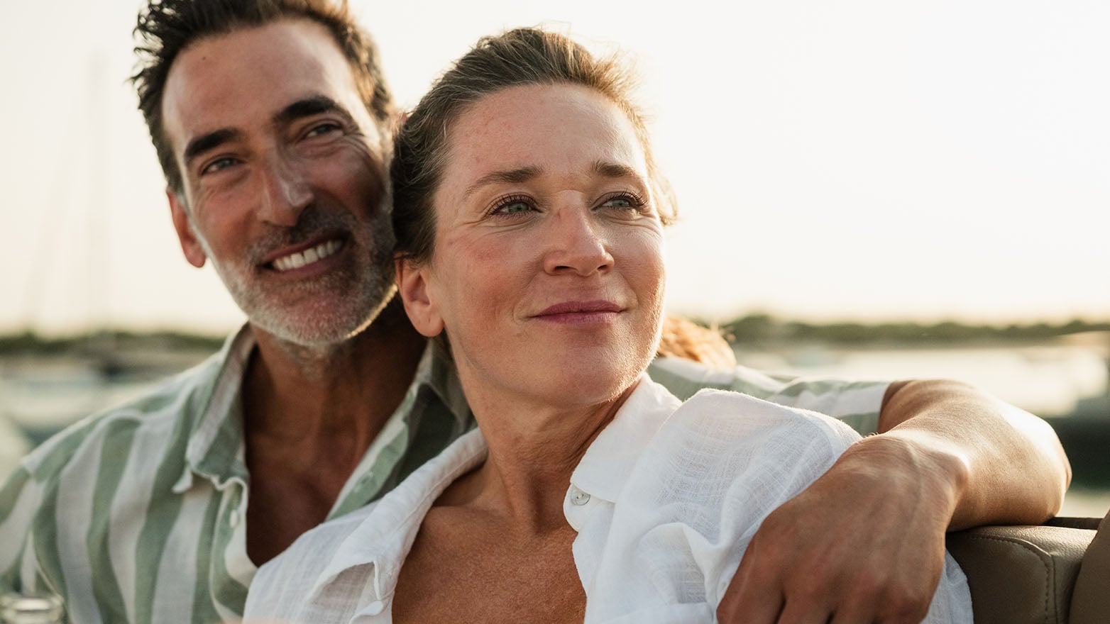 Two people sit close together on a boat, with one person’s arm resting along the back of the seat as water and shoreline appear in the background.