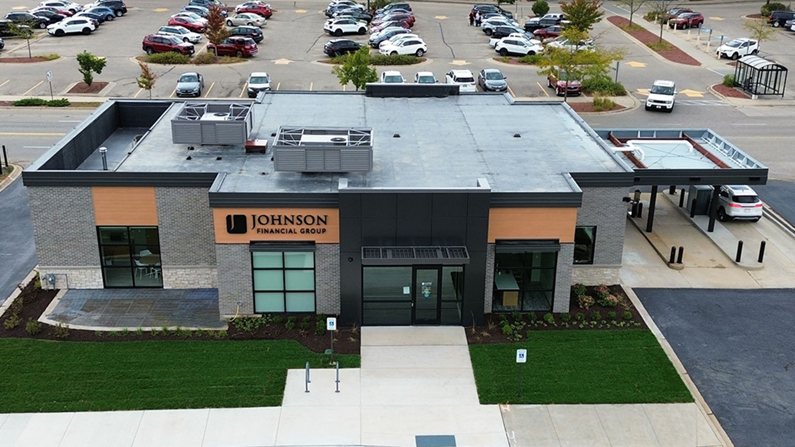 An eye-level photograph captures a modern brick building housing the "Johnson Financial Group," surrounded by a vibrant green lawn. The building features large, multi-paned windows flanking the entrance, and a drive-through service on one side. In the background, a parking lot is filled with numerous vehicles.