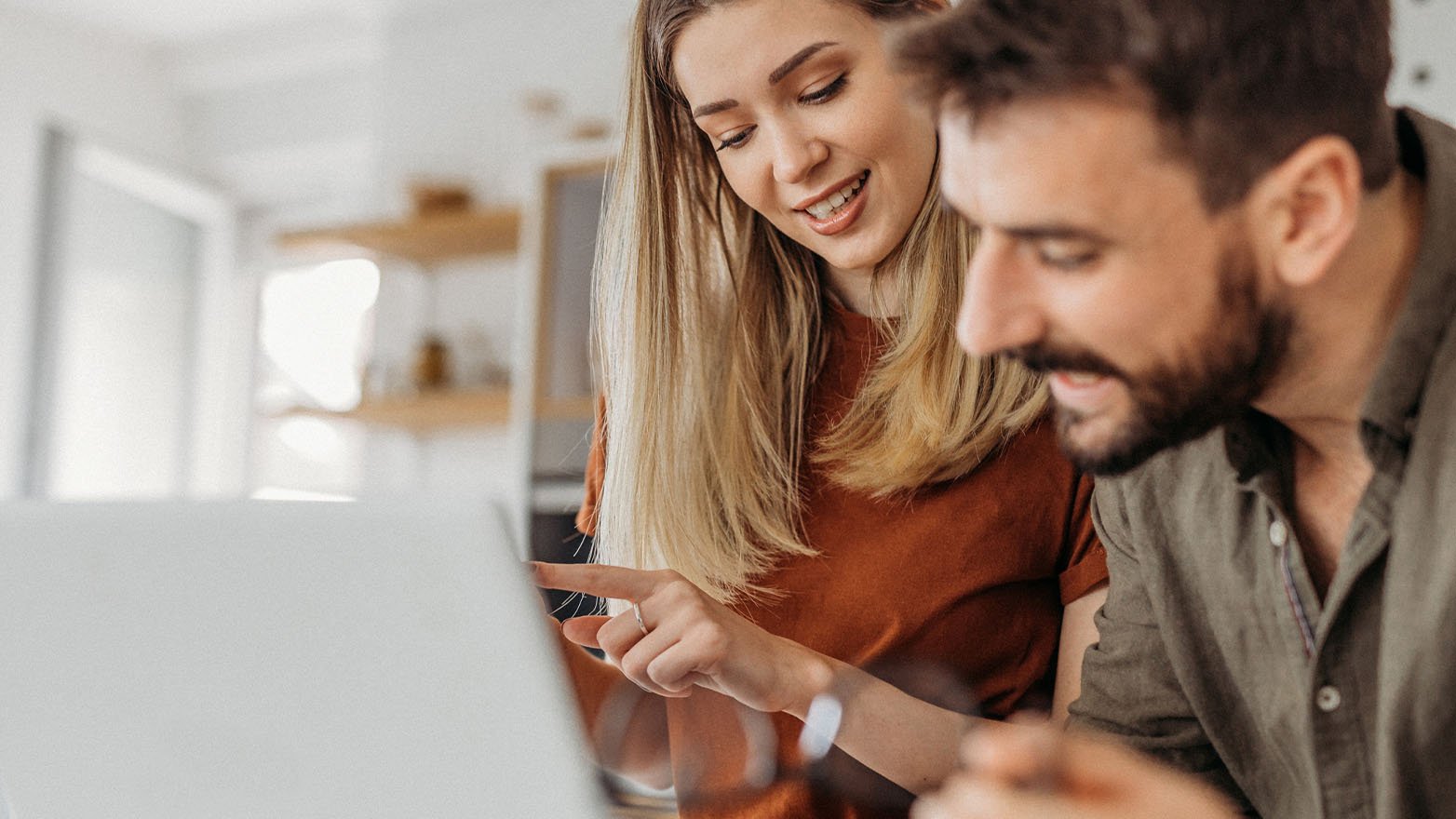A smiling woman points at a laptop screen shared with a man.