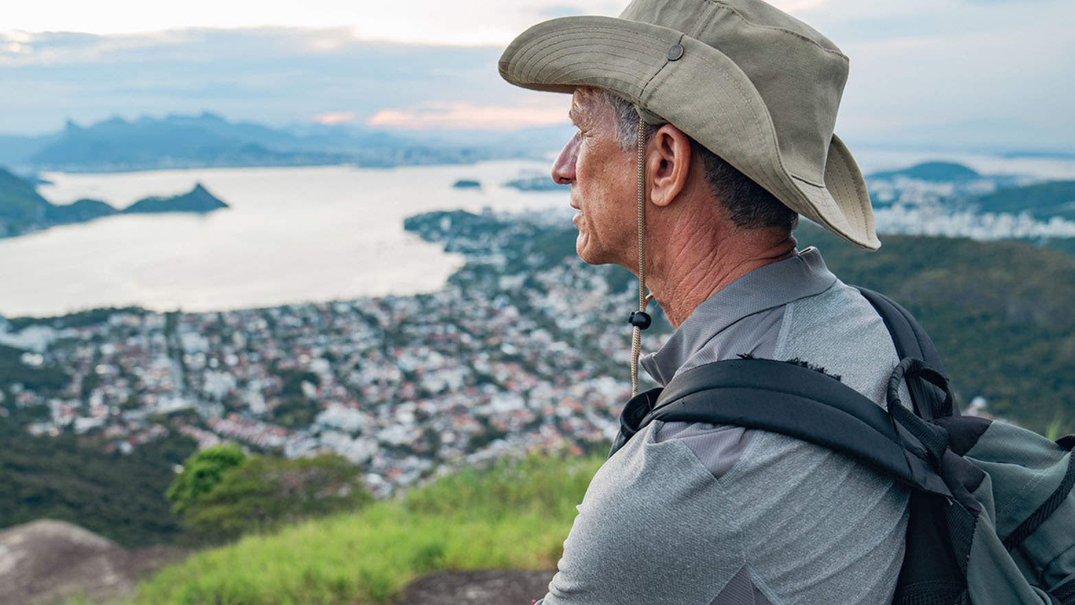 A hiker with a backpack stands on a grassy overlook, taking in a sweeping view of a coastal town and water below.