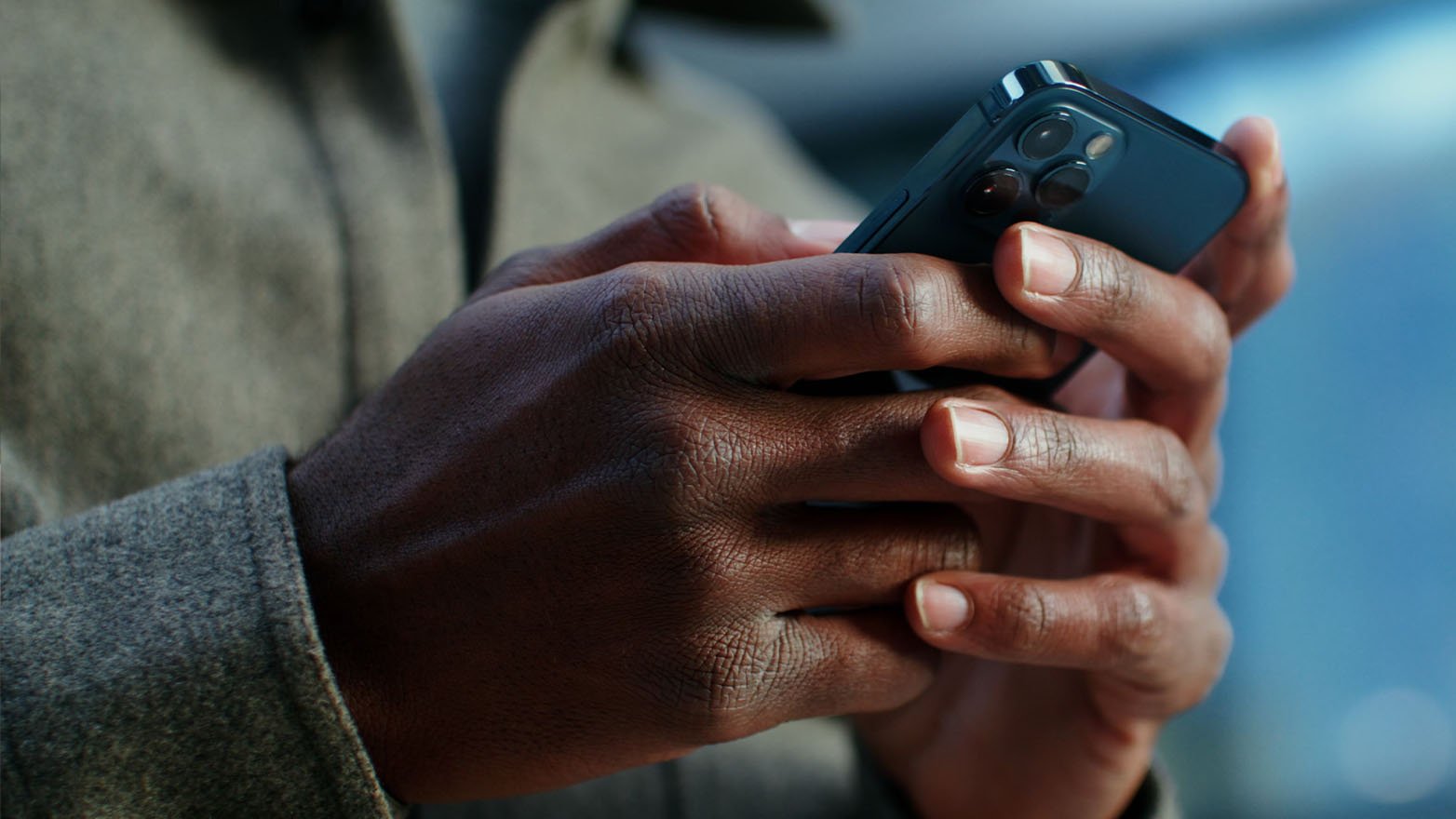 Close-up of dark-skinned hands holding a blue iPhone with a triple-lens camera. The person is wearing a gray woolen coat.