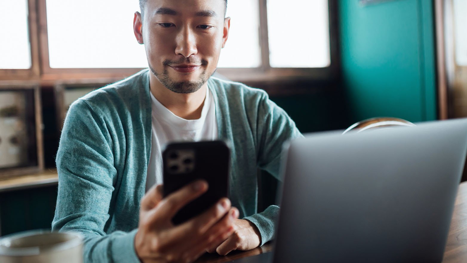 A person sits at a café table, holding a smartphone while working on a laptop.