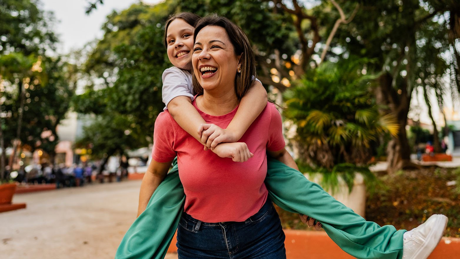 An adult carries a child on their back while walking along a path in a green park.