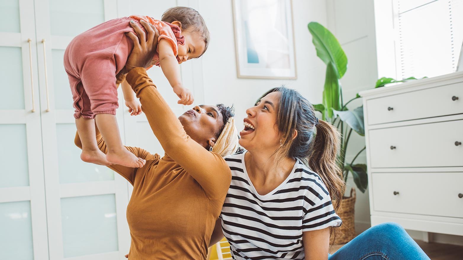A woman holds a baby above her head while another woman sits next to her, looking up at the baby and smiling. The woman holding the baby wears a brown long-sleeved shirt and blue jeans and has her arms extended straight up to hold the baby. The baby wears a pink onesie and is looking down at the woman holding her. The woman next to them wears a black-and-white striped shirt and blue jeans and has her mouth open in a smile. They are all sitting on a colorful rug in a room with white walls and a white dresser.