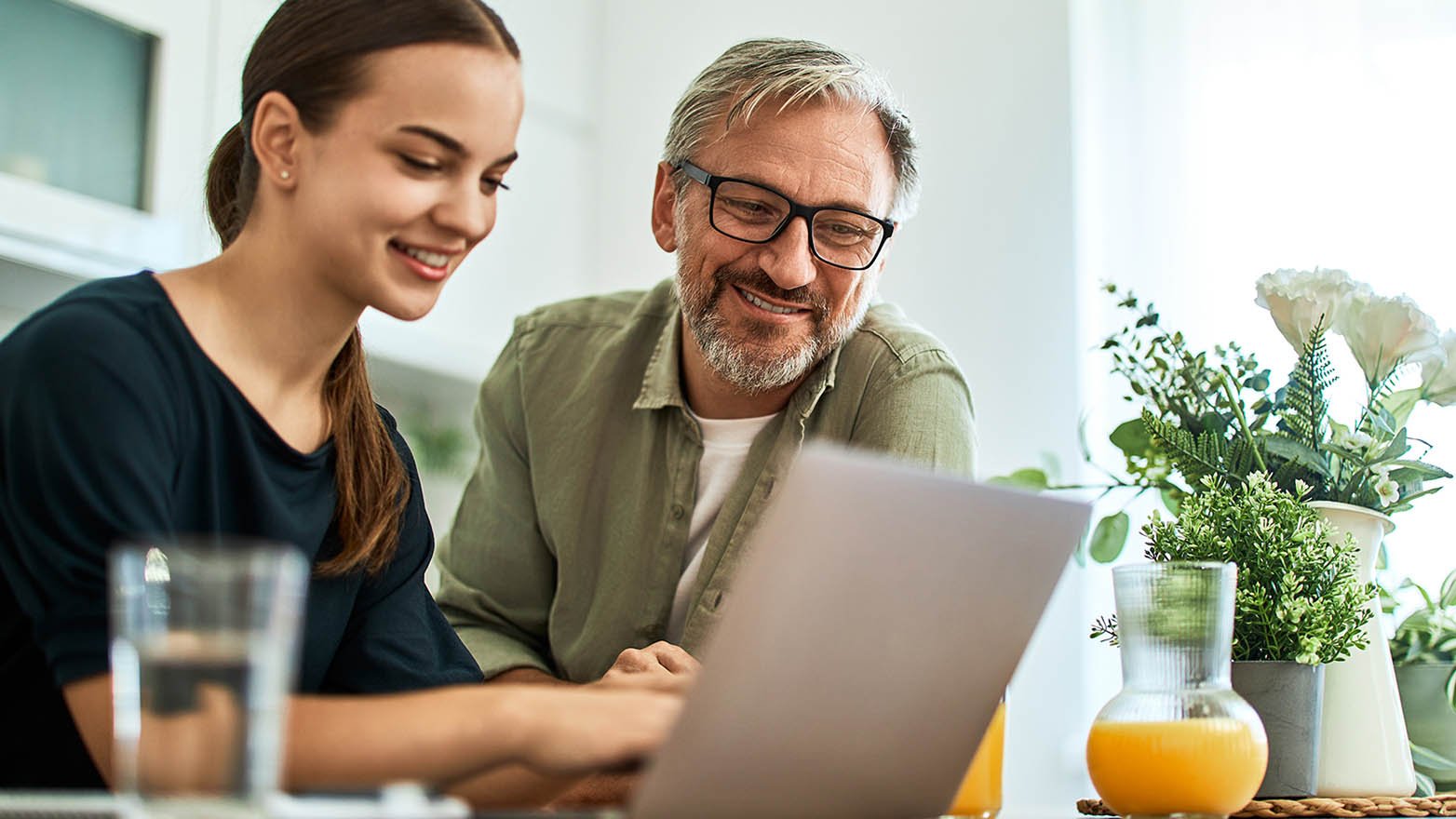 A smiling young woman and an older man, possibly her father, are sitting at a table looking at a laptop screen. The woman is typing on the laptop, and the man is watching the screen with a smile. There are drinks and plants on the table.