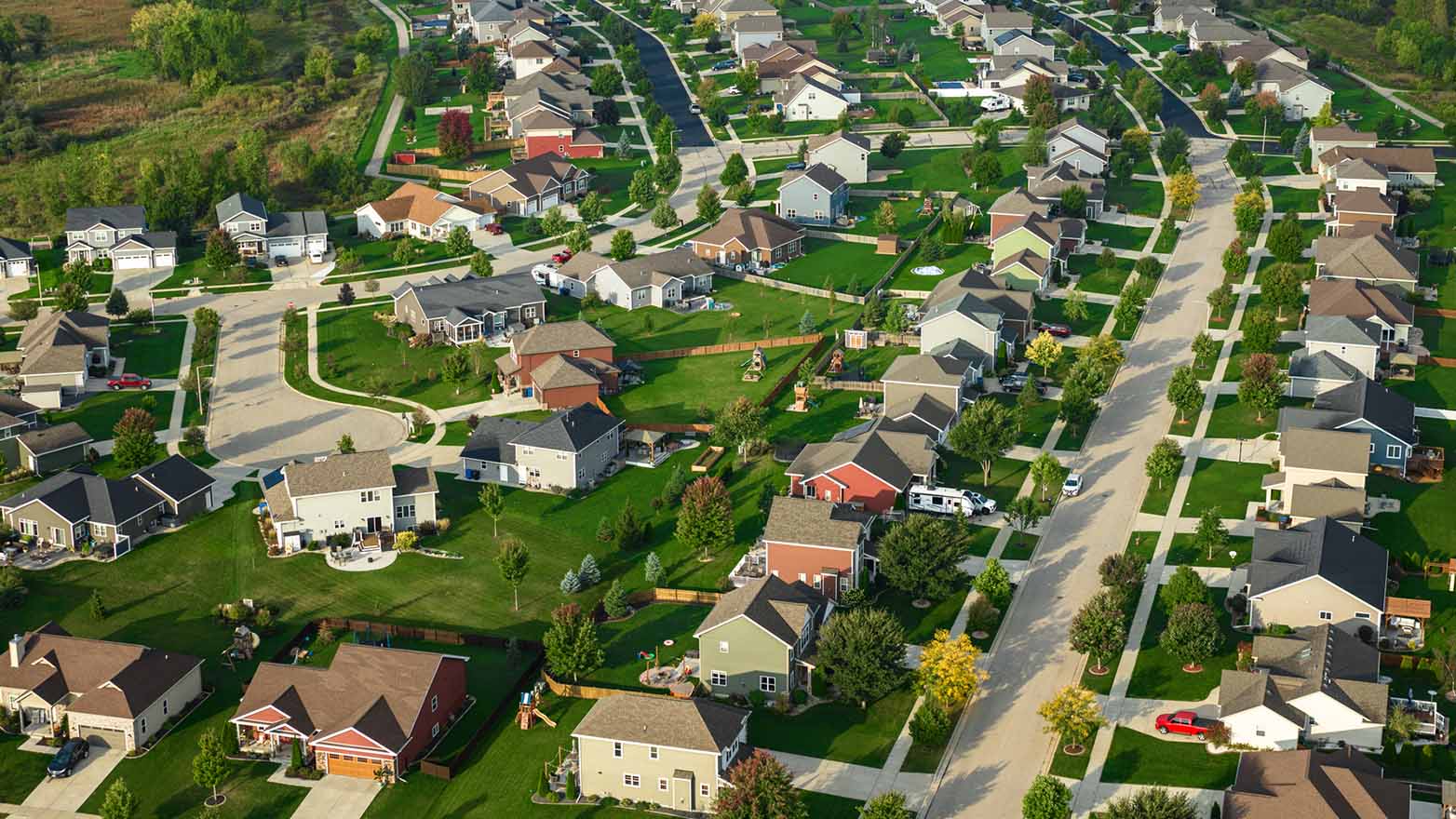 Aerial view of a suburban neighborhood with rows of single-family homes, tree-lined streets, sidewalks, and well-maintained green lawns. Houses are arranged along curved residential roads with spacious backyards and scattered vehicles visible throughout the community.