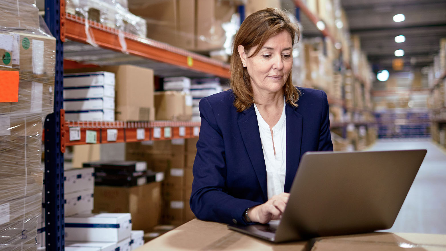 Professional woman in navy blazer using laptop in warehouse facility with orange shelving and inventory boxes in background