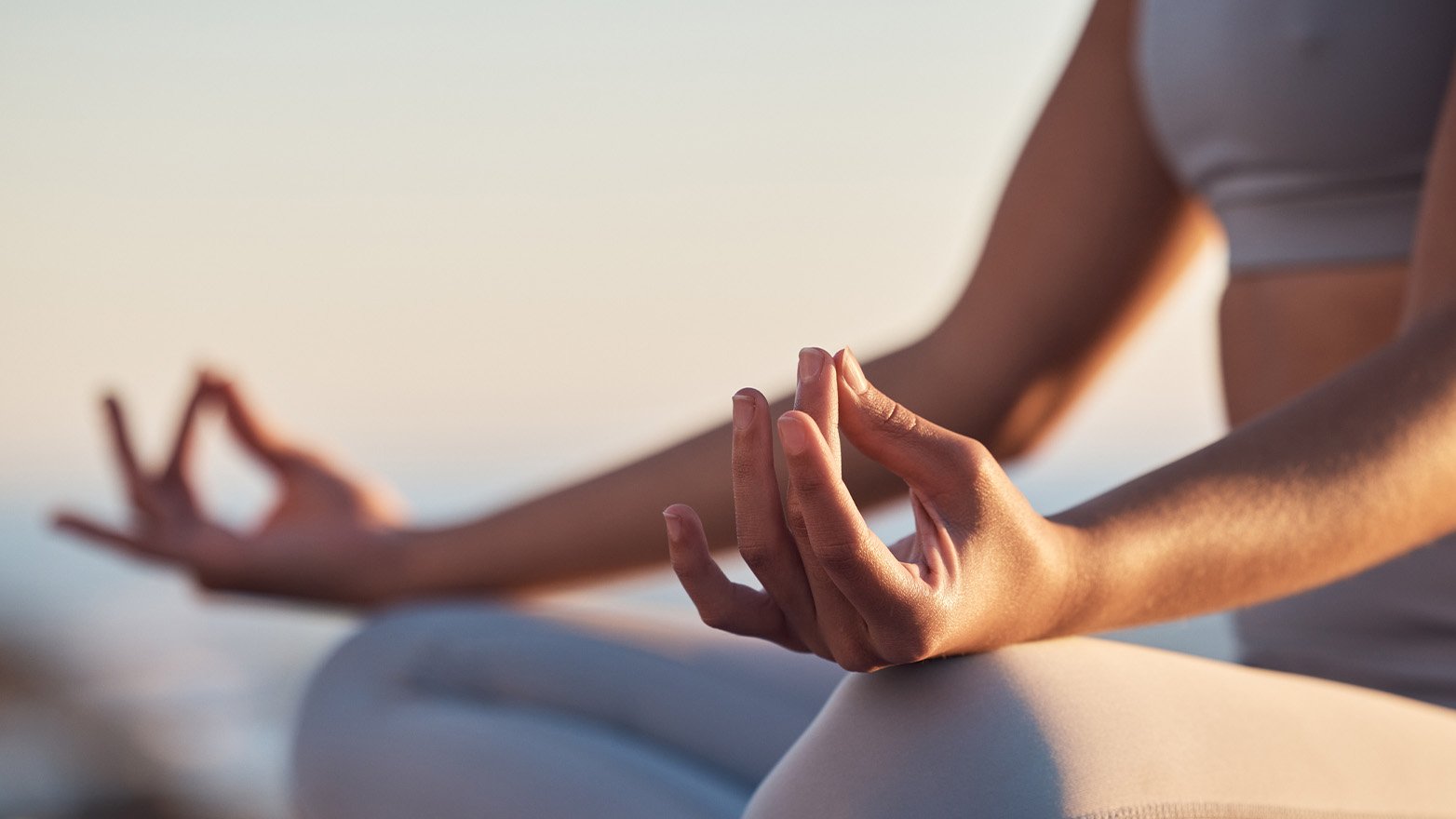 A close up image of a woman's torso and arms as she sits cross legged in meditation. Her hands are resting on her knees in the Gyan Mudra, with the tips of her thumbs and index fingers touching. She is wearing light gray athletic wear and the background is a soft, hazy light.