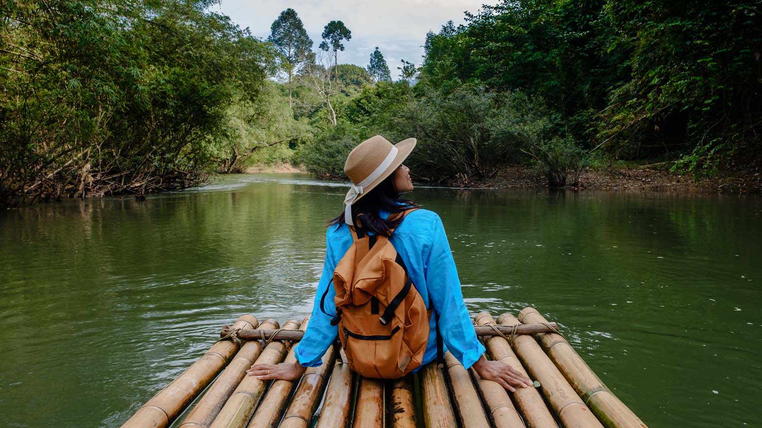 A person wearing a sun hat and backpack sits on a bamboo raft floating on a calm river, surrounded by lush green trees and vegetation. The person faces the water and forest ahead, creating a peaceful outdoor scene.