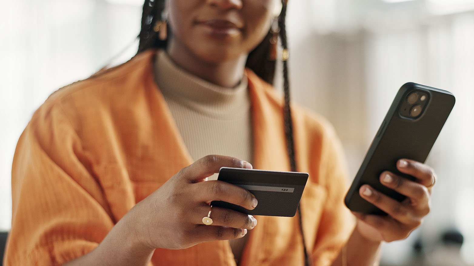 Close-up of a woman in an orange shirt holding a smartphone and credit card, appearing to make an online purchase.
