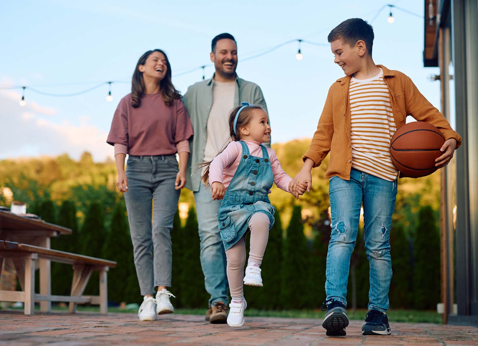 Family of four walking outside together at sunset, with an older boy holding a basketball and holding hands with a young girl while their parents smile behind them.