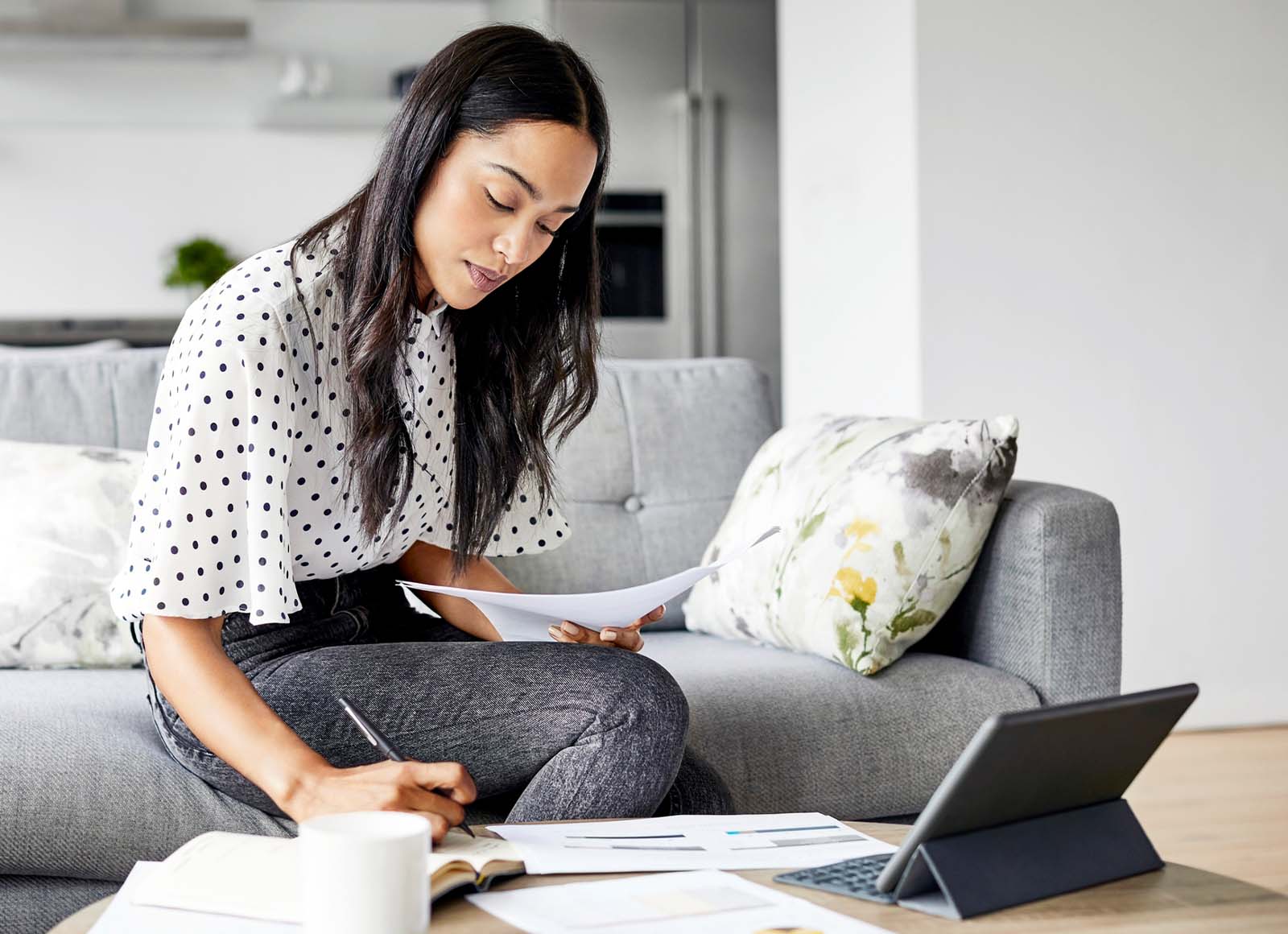 Woman in polka dot blouse sitting on gray couch, holding papers while using calculator with laptop open on coffee table in modern home setting