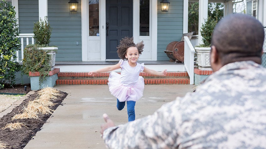 Dad dressed in fatigues bending down to greet his daughter in the front yard.