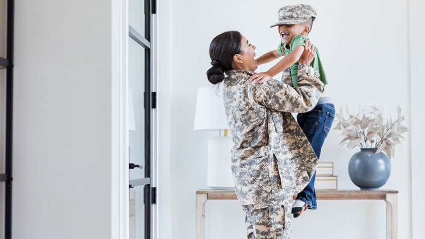 Mom dressed in military fatigues lifting her son in the air in her living room.