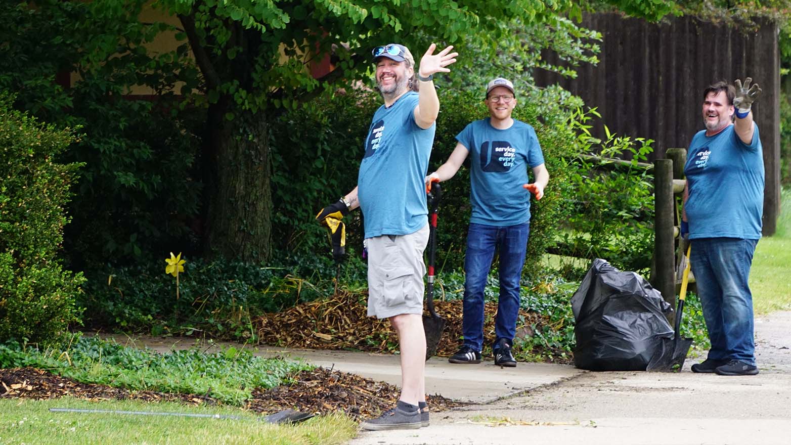 Three men, dressed alike in blue t-shirts and gloves, stand outdoors near a large black trash bag. The man on the left waves his arm in greeting.