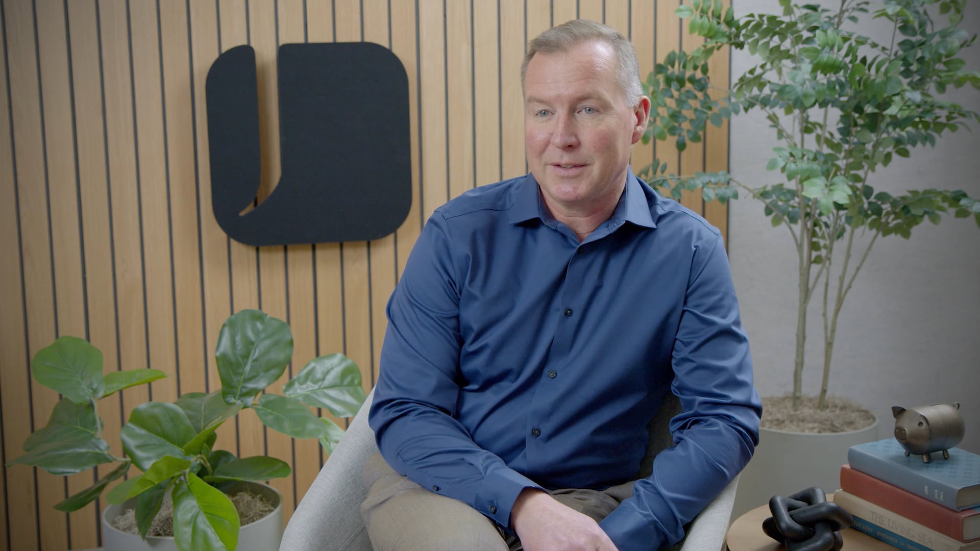 A man in a blue button-down shirt sits in an armchair in a modern office, in front of a wood-paneled wall with a large black logo.