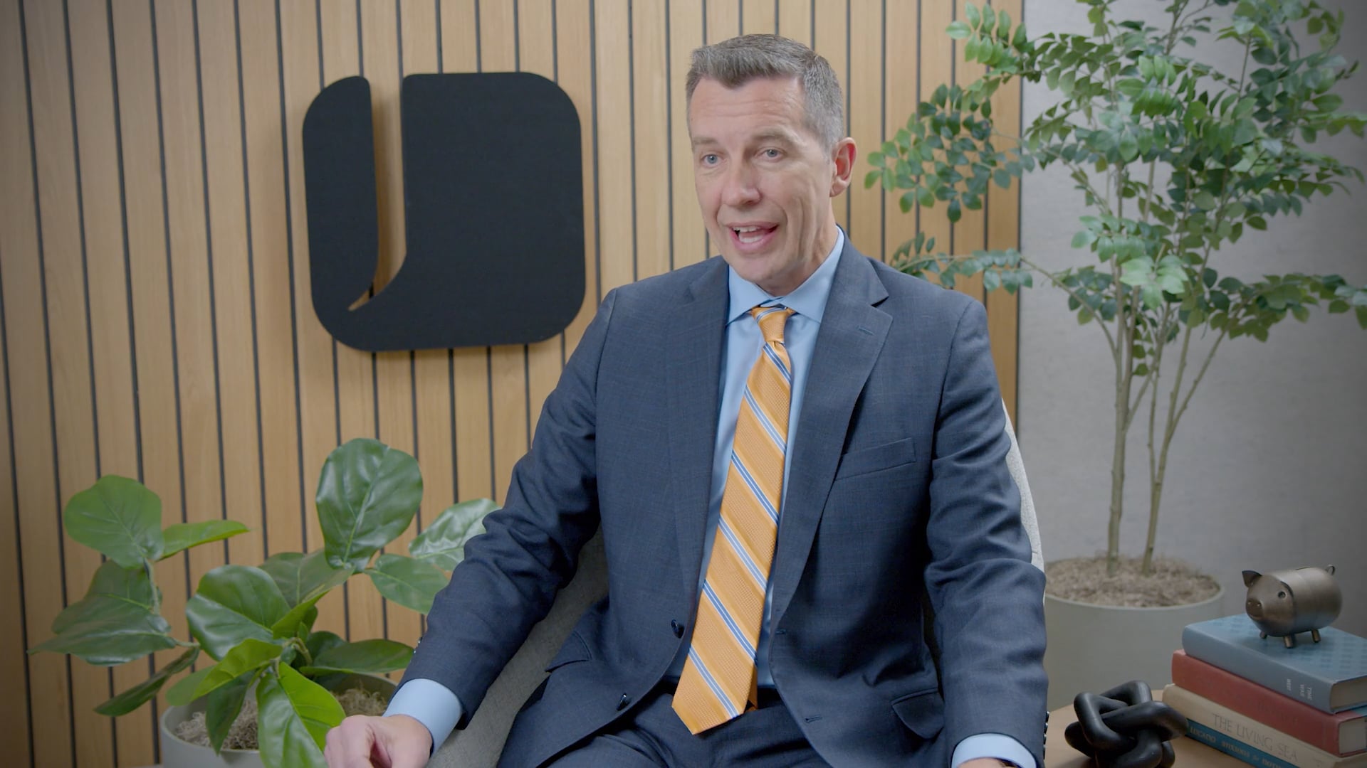 A person wearing a dark suit with a yellow striped tie sits in a modern office chair. Behind the person is a wood slat wall with a black abstract logo, along with green potted plants.