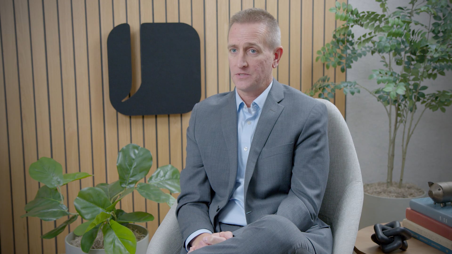 A man in a grey suit and light blue shirt sits in an armchair in a modern office. Behind him, a wood-paneled wall features a black speech-bubble logo and is flanked by potted plants.