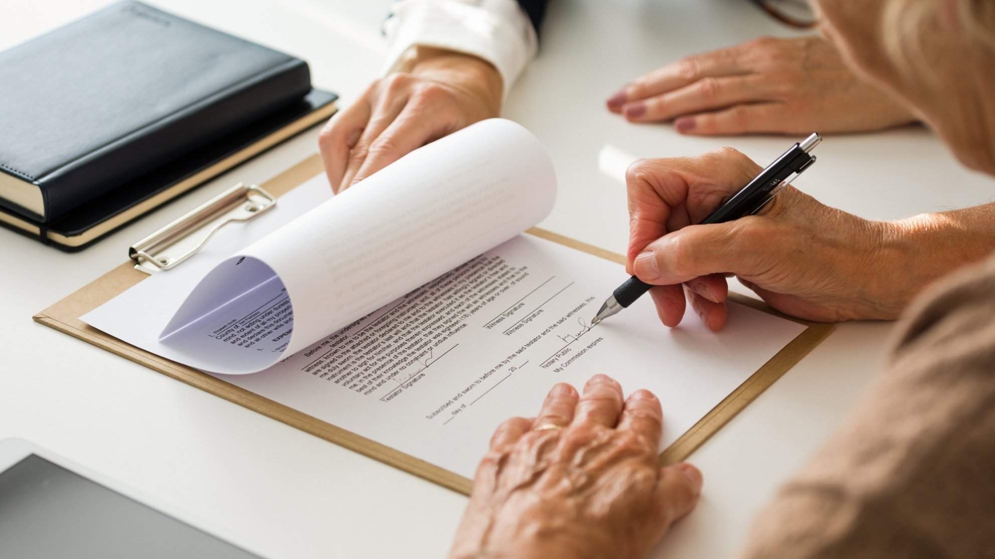 A person signs a document on a clipboard, closely observed by a witness, in a close-up view from directly above.