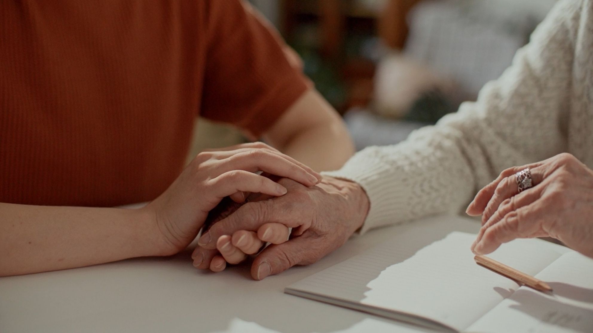 A younger person's hands supportively hold the hands of an older person, who sits at a table with a notebook and pencil.