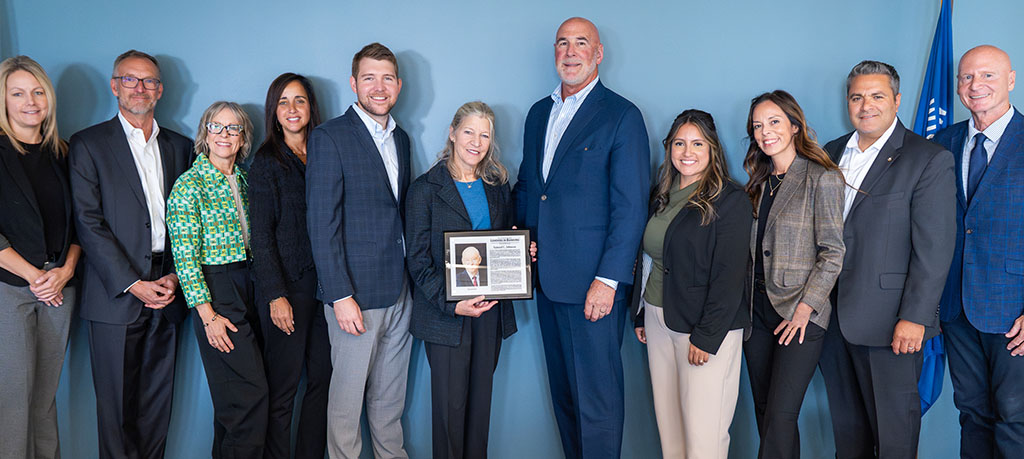 Group of twelve people standing in a line indoors against a blue wall, with U.S. and Wisconsin flags visible at either side; one person near the center is holding a framed plaque.